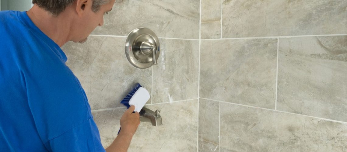 A man doing household chores is cleaning bathroom bathtub tiles and fixtures with a soapy white scrub brush.